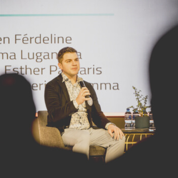 Man presenting on stage with microphone, audience silhouettes in foreground.