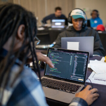 Close-up of a laptop screen displaying code, with a blurred user in the foreground and other hackathon participants working on laptops in the background.